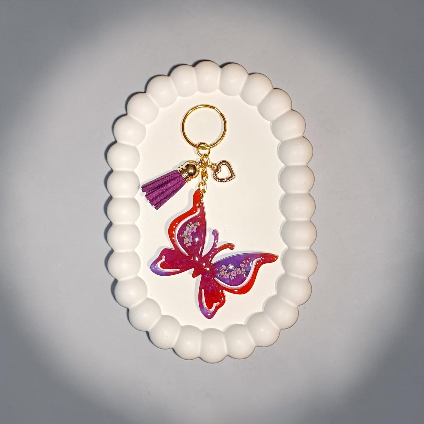 Portachiavi farfalla Lunéa in resina arancione e viola con dettagli floreali, charm dorati e nappina porpora esposto su vassoio decorativo bianco.
